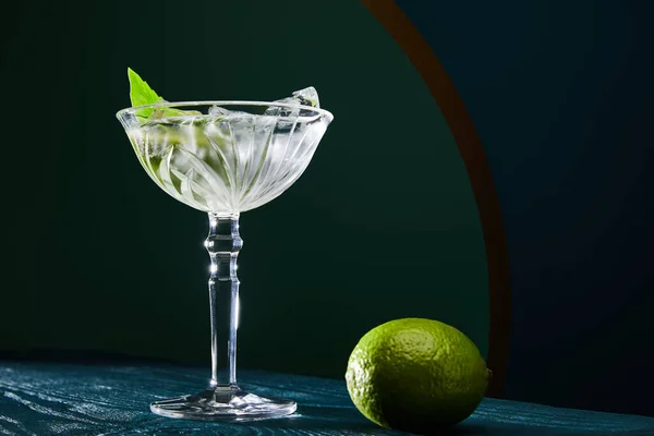 Vista a basso angolo del bicchiere da cocktail con cubetti di ghiaccio, foglia di menta e calce intera su una superficie di legno blu su sfondo geometrico — Foto stock