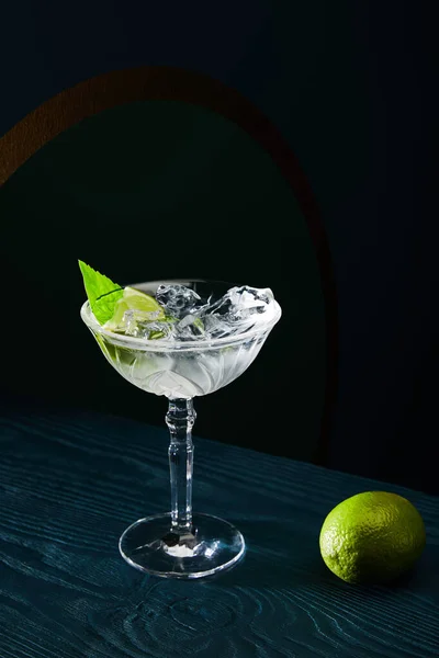 Hochwinkelblick auf Cocktailglas mit Eiswürfeln, Minzblatt und ganzer Limette auf blauer Holzoberfläche auf schwarzem Hintergrund — Stockfoto