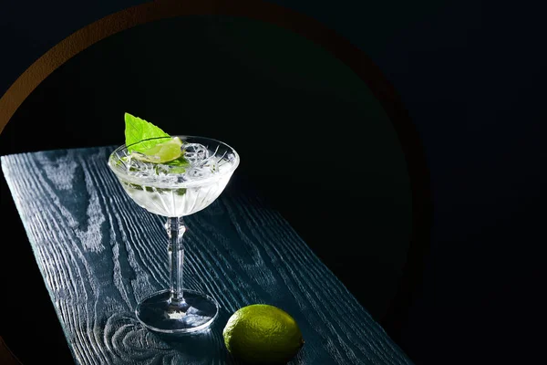 Vue grand angle du verre à cocktail avec boisson avec glaçons, feuille de menthe et citron vert entier sur une surface en bois bleu sur fond noir — Photo de stock