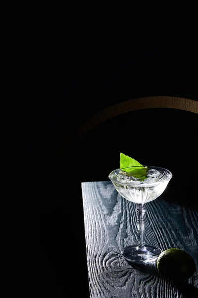 Vista de ángulo alto de copa de cóctel con bebida con cubitos de hielo, hoja de menta y lima entera sobre superficie de madera azul sobre fondo negro con líneas geométricas - foto de stock