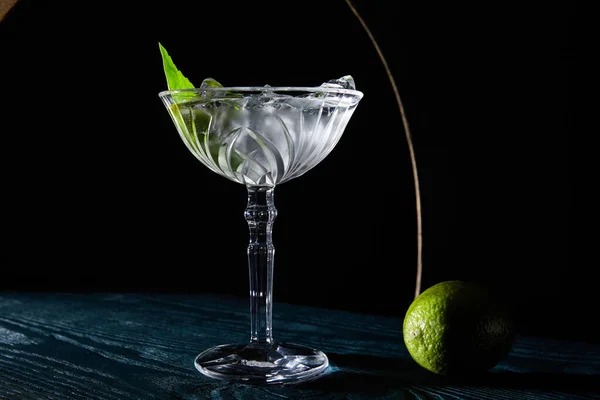 Bicchiere da cocktail con bevanda con cubetti di ghiaccio, foglia di menta e lime intero su superficie di legno blu su fondo nero con linee geometriche — Foto stock