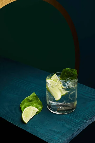 Vue grand angle du verre à l'ancienne avec boisson fraîche, feuille de menthe et tranche de citron vert sur une surface en bois bleu sur fond géométrique vert et bleu — Photo de stock