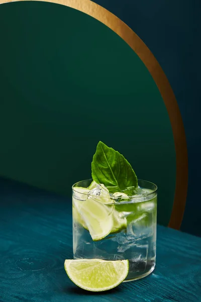 Old fashioned glass with fresh drink, mint leaf and lime slice on blue wooden surface on green and blue geometric background with golden lines — Stock Photo