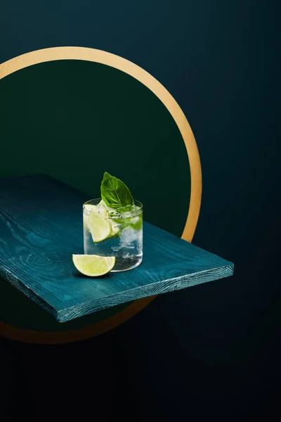 Vista ad alto angolo di vetro vecchio stile con bevanda fresca, foglia di menta e fetta di lime su superficie di legno blu su sfondo geometrico verde e blu con cerchio dorato — Foto stock