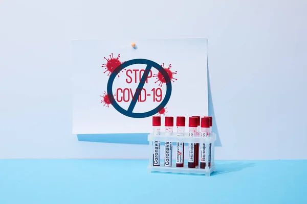 Paper with stop covid-19 lettering near test tubes with coronavirus test on blue — Stock Photo