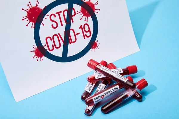Papier mit Stop Covid-19 Schriftzug in der Nähe von Reagenzgläsern mit Blutproben auf blau — Stockfoto