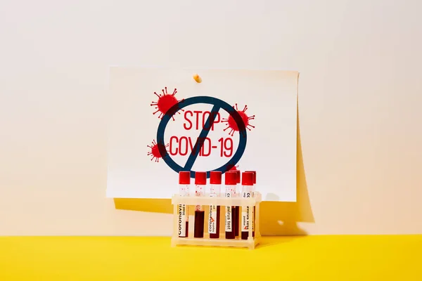 Paper with stop covid-19 lettering near test tubes with coronavirus test on white and yellow — Stock Photo