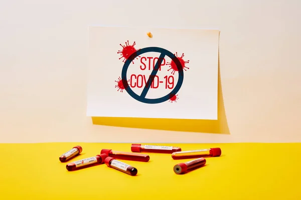 Papel con letras stop covid-19 cerca de los tubos de ensayo con muestras de sangre en blanco y amarillo - foto de stock