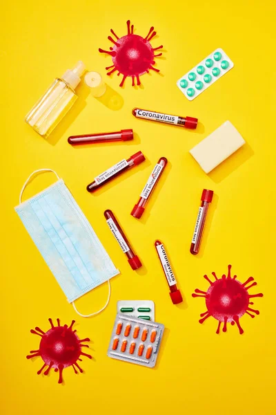 Top view of test tubes with coronavirus lettering near medical mask, blister packs with pills, bottle with hand sanitizer and soap bar on yellow — Stock Photo