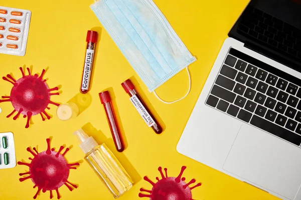 Top view of test tubes with coronavirus lettering near medical mask, blister packs with pills, bottle with antibacterial liquid and laptop on yellow — Stock Photo