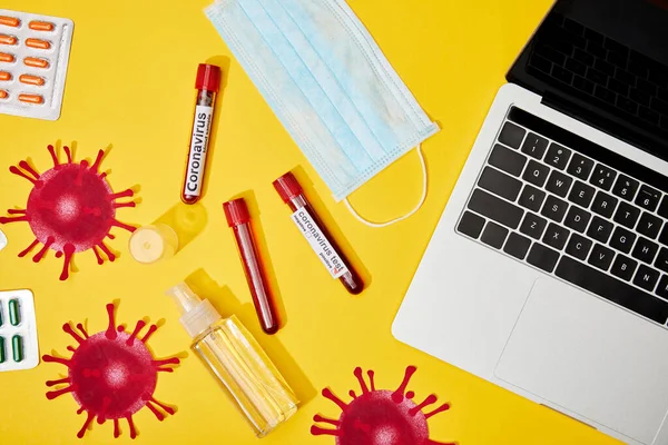 Вид пробирки с коронавирусом рядом с медицинской маской, таблетки, бутылка с антибактериальной жидкостью и ноутбук на желтом — стоковое фото