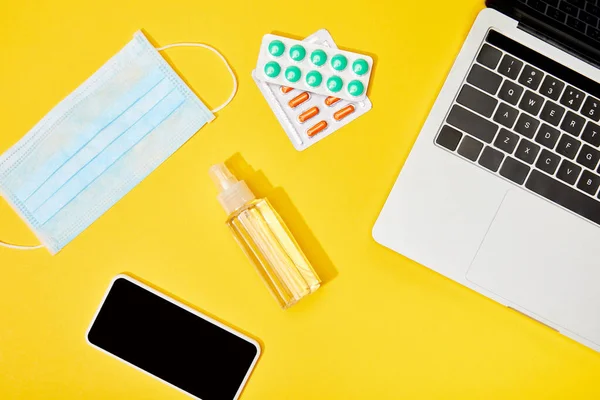 Vista superior do laptop e smartphone com tela em branco perto de pílulas, máscara médica e garrafa com desinfetante de mão no amarelo — Fotografia de Stock