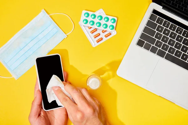 Vista superior del hombre que sostiene la servilleta cerca del teléfono inteligente con la pantalla en blanco, ordenador portátil, píldoras, desinfectante de manos y máscara médica en amarillo - foto de stock