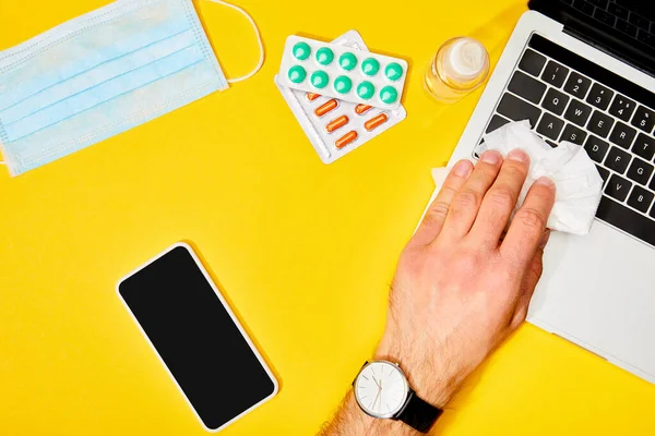 Top view of man holding napkin near laptop, smartphone with blank screen, pills, hand sanitizer and medical mask on yellow — Stock Photo