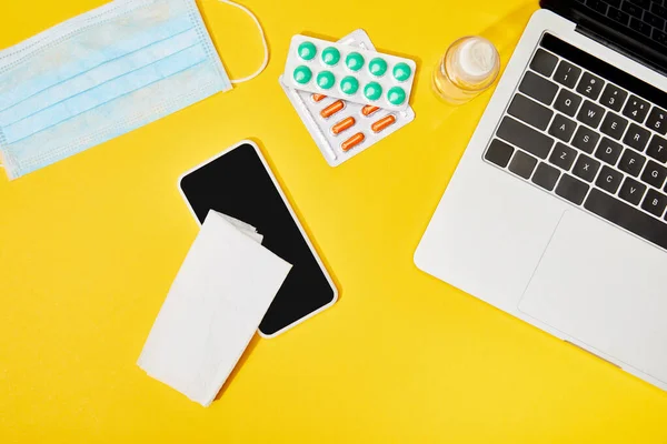 Вид сверху ноутбука и смартфона с чистым экраном рядом с салфеткой, таблетки, медицинская маска и бутылка с дезинфицирующим средством для рук на желтом — стоковое фото