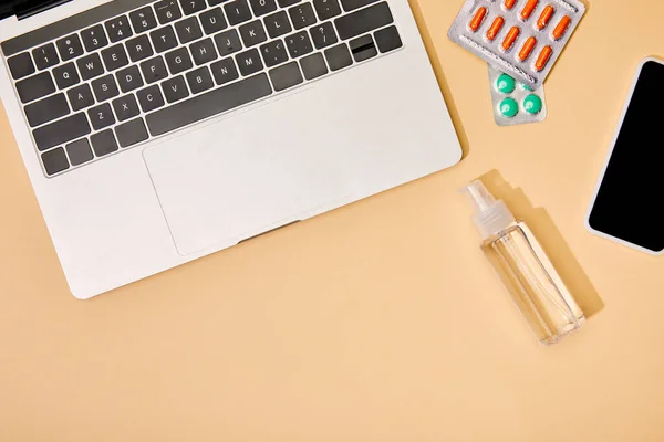 Vista superior de la computadora portátil y el teléfono inteligente con pantalla en blanco cerca de píldoras y botella con desinfectante de manos en beige - foto de stock