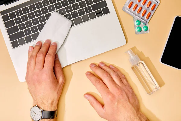 Vue du dessus de l'homme tenant la serviette près de l'ordinateur portable, smartphone avec écran blanc, pilules et désinfectant pour les mains sur beige — Photo de stock