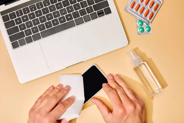 Vista superior do homem segurando guardanapo perto do smartphone com tela em branco, laptop, pílulas e desinfetante de mão no bege — Fotografia de Stock