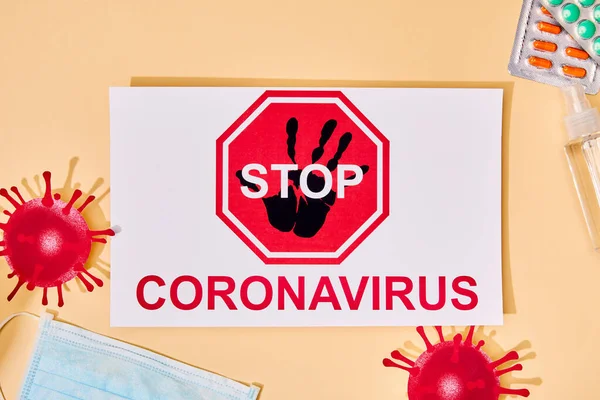 Верхний вид бумаги с остановкой коронавирусной надписи рядом с нарисованным вирусом, медицинская маска, дезинфицирующее средство для рук и таблетки на бежевом — стоковое фото