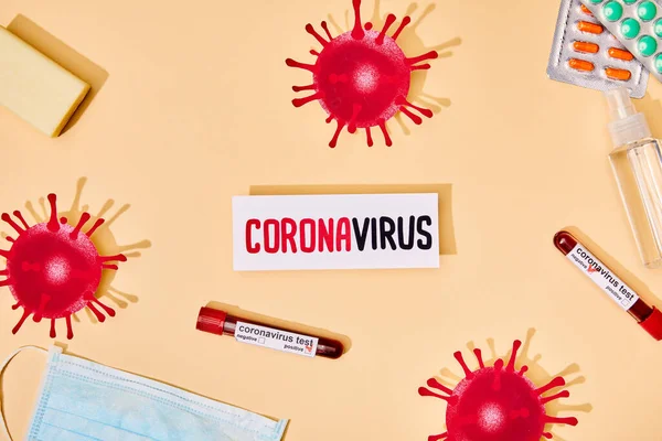 Top view of paper with coronavirus lettering near drawn virus, test tubes, soap bar, medical mask, pills and hand sanitizer on beige — Stock Photo