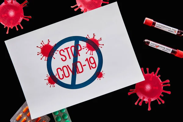 Top view of drawn virus near paper with stop covid-19 lettering, pills and test tubes with coronavirus test isolated on black — Stock Photo