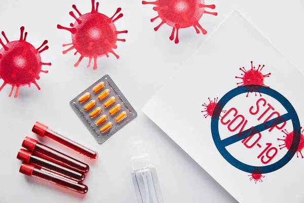 Top view of placard with stop covid-19 lettering near drawn virus, test tubes, hand sanitizer and pills on white — Stock Photo