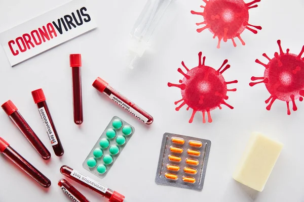 Vista dall'alto di carta con scritte coronavirus vicino a virus disegnato, provette, disinfettante per le mani, sapone bar e pillole su bianco — Foto stock