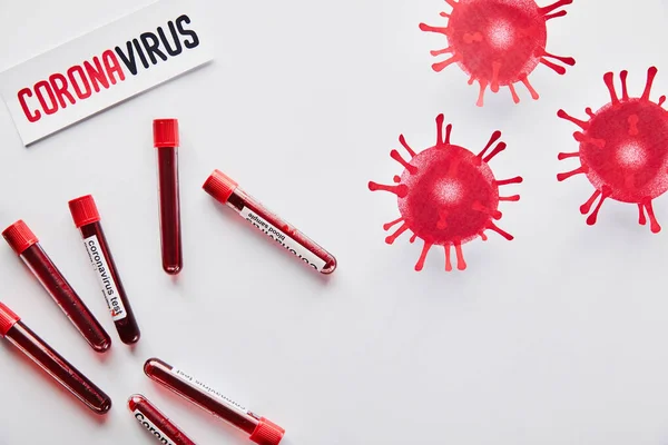 Vue du dessus des éprouvettes avec des échantillons de sang et test de coronavirus près de virus dessinés et du papier avec lettrage de coronavirus sur blanc — Photo de stock