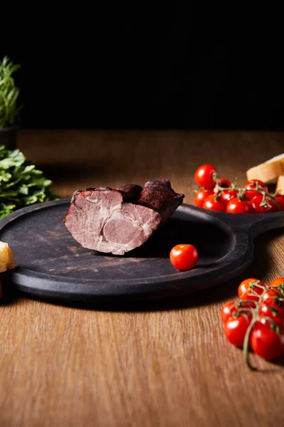 Foco seletivo de presunto saboroso a bordo perto de salsa, tomates cereja e baguete em mesa de madeira isolada em preto — Stock Photo
