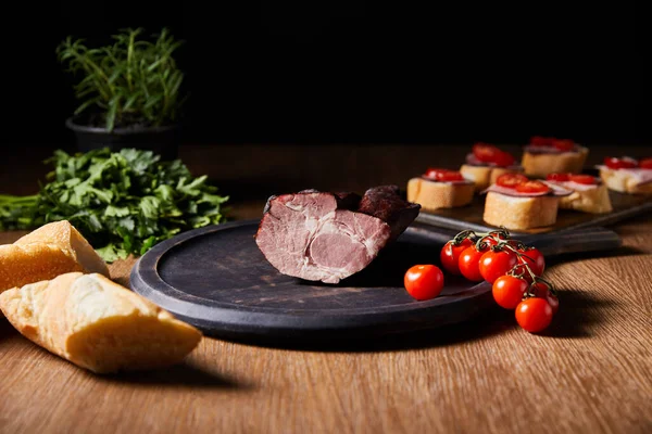 Foyer sélectif de jambon savoureux à bord près de canape, persil, tomates cerises et baguette sur table en bois isolé sur noir — Photo de stock