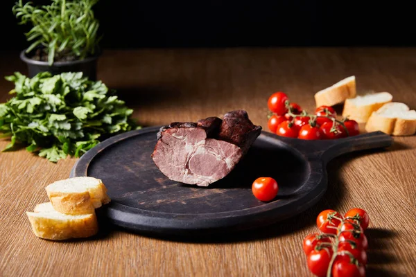 Enfoque selectivo de sabroso jamón a bordo cerca de perejil, tomates cherry y baguette en mesa de madera aislado en negro - foto de stock