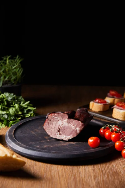 Selective focus of tasty ham on board near canape, parsley, cherry tomatoes and baguette on wooden table isolated on black — Stock Photo