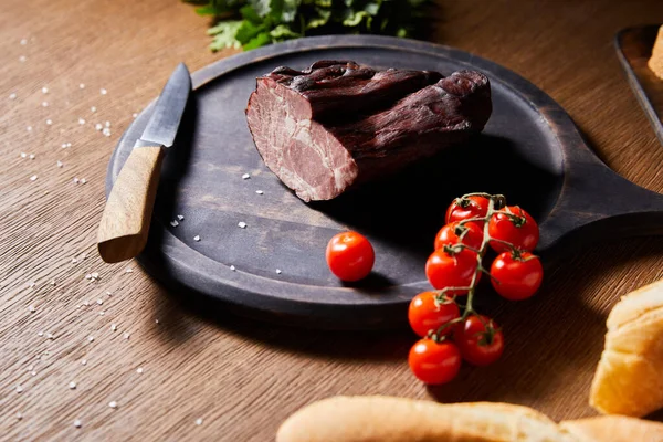 Enfoque selectivo de sabroso jamón a bordo cerca del perejil, tomates cherry y baguette con sal dispersa y cuchillo en la mesa de madera - foto de stock