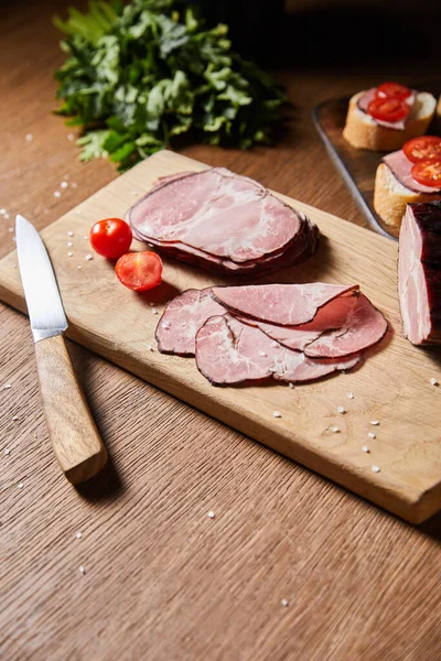 Selective focus of tasty ham slices, cherry tomatoes and knife on cutting board near parsley and canape — Stock Photo