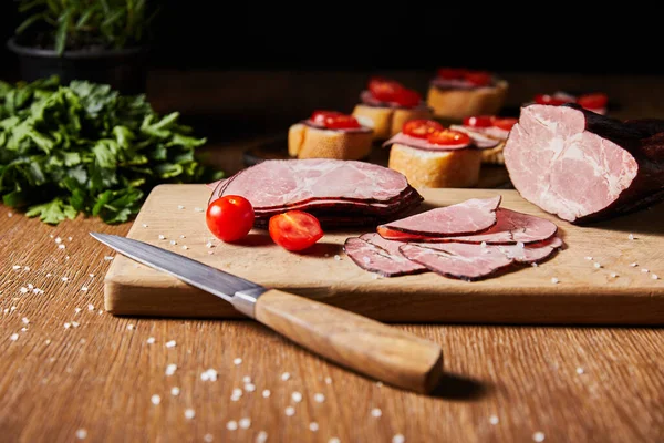 Foco seletivo de fatias de presunto saborosas, tomates cereja e faca na tábua de corte perto de salsa e canapé — Fotografia de Stock