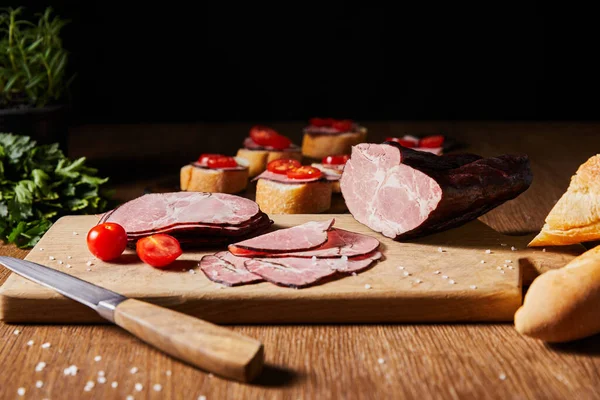 Selective focus of tasty ham slices, cherry tomatoes and knife on cutting board near parsley and canape — Stock Photo