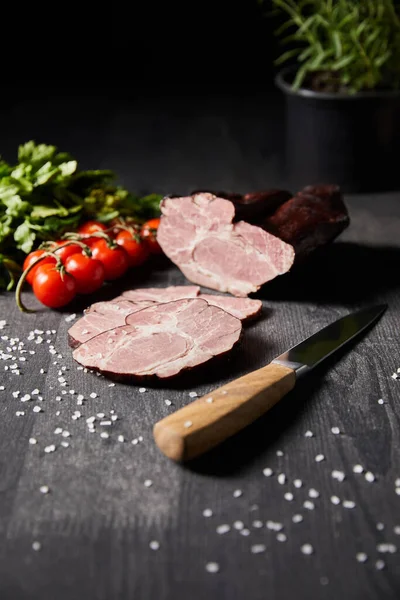 Fuoco selettivo di prosciutto affettato saporito, pomodorini, prezzemolo, sale, coltello su tavola grigia di legno — Foto stock