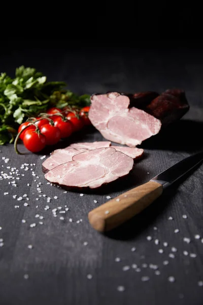 Selective focus of tasty ham sliced ham, cherry tomatoes, parsley, salt, knife on wooden grey table isolated on black — Stock Photo