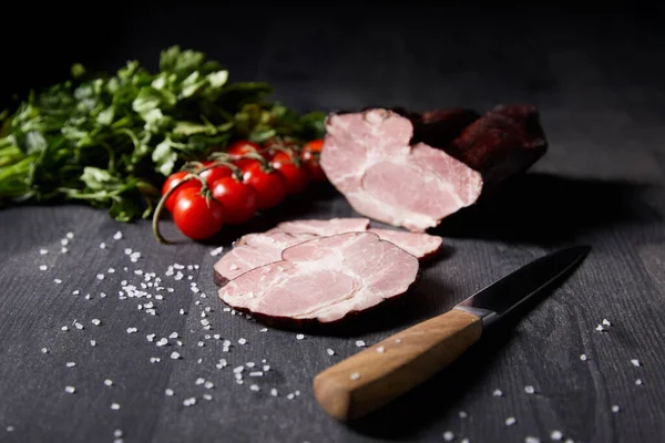 Foco seletivo de presunto saboroso presunto fatiado, tomate cereja, salsa, sal, faca na mesa cinza de madeira — Fotografia de Stock