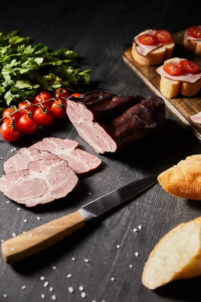 Selective focus of tasty ham sliced ham, cherry tomatoes, parsley, salt, knife and baguette on wooden grey table with canape — Stock Photo
