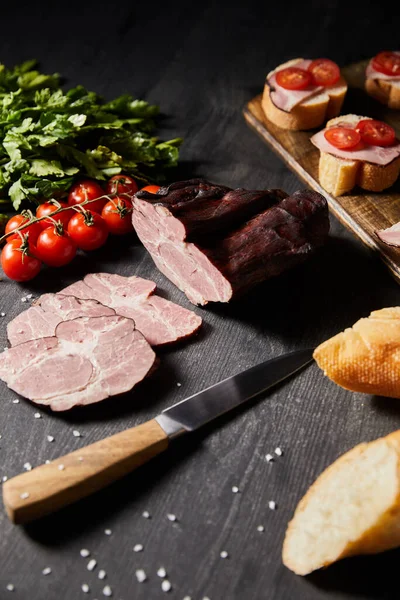 Focus selettivo di gustoso prosciutto affettato, pomodorini, prezzemolo, sale, coltello e baguette su tavola grigia in legno con tartina — Foto stock