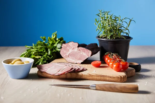 Selective focus of tasty ham on cutting board with knife, parsley, cherry tomatoes, olives on wooden table on blue background — Stock Photo