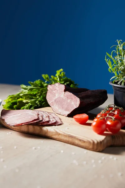Fuoco selettivo di prosciutto saporito su tagliere con prezzemolo, pomodorini su tavolo di legno su sfondo blu — Foto stock