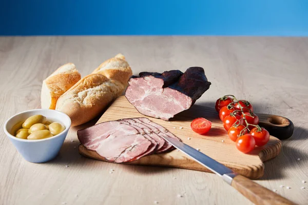 Tasty ham on cutting board with knife, cherry tomatoes, olives and baguette on wooden table on blue background — Stock Photo