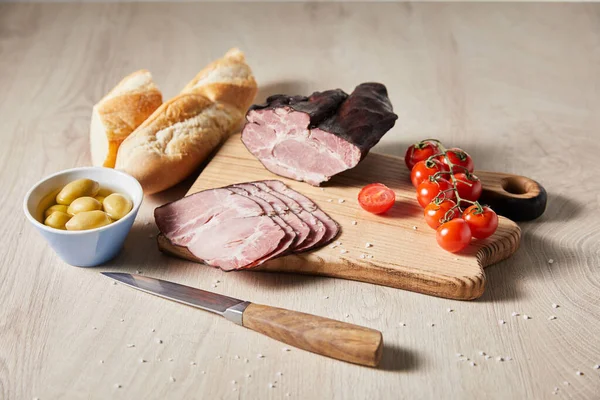 Selettiva messa a fuoco del gustoso prosciutto sul tagliere con coltello, pomodorini, olive e baguette sul tavolo di legno — Foto stock