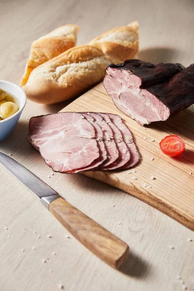 Selective focus of tasty ham on cutting board with knife, cherry tomato, olives and baguette on wooden table — Stock Photo