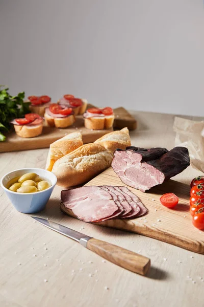 Selective focus of tasty ham on cutting board with knife, parsley, cherry tomatoes, olives and baguette near canape on wooden table isolated on grey — Stock Photo