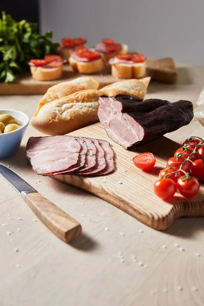 Selective focus of tasty ham on cutting board with knife, parsley, cherry tomatoes, olives and baguette near canape on wooden table — Stock Photo