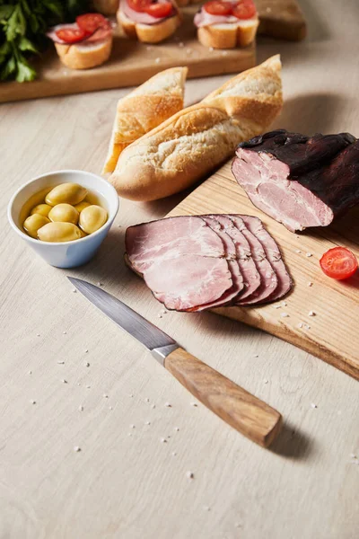 Enfoque selectivo de sabroso jamón en la tabla de cortar con cuchillo, aceitunas y baguette en la mesa de madera - foto de stock