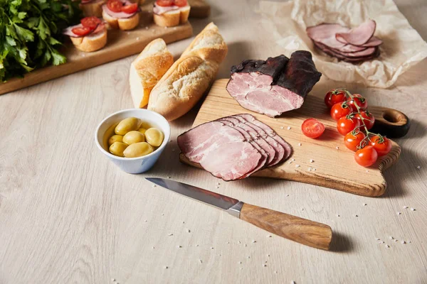 Selective focus of tasty ham on cutting board with knife, parsley, cherry tomatoes, olives and baguette near canape on wooden table — Stock Photo